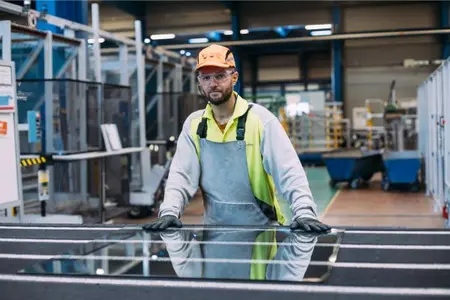 Pracovník ve výrobním provozu Glassolutions stojí u velké tabule skla na pracovním stole. Je vybaven ochrannou vestou, rukavicemi a sluchátky. Okolo něj jsou vidět výrobní stroje, kovové konstrukce a průmyslové vybavení haly.