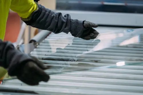 Pracovník v ochranných rukavicích manipuluje s velkým skleněným panelem na válečkové lince ve výrobním provozu.