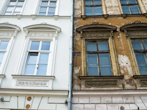 Detail fasády dvou sousedních domů – vlevo renovovaná světlá budova s novými okny a dekorativními prvky, vpravo starší, opotřebená fasáda se zašlými rámy oken a poškozenou omítkou.